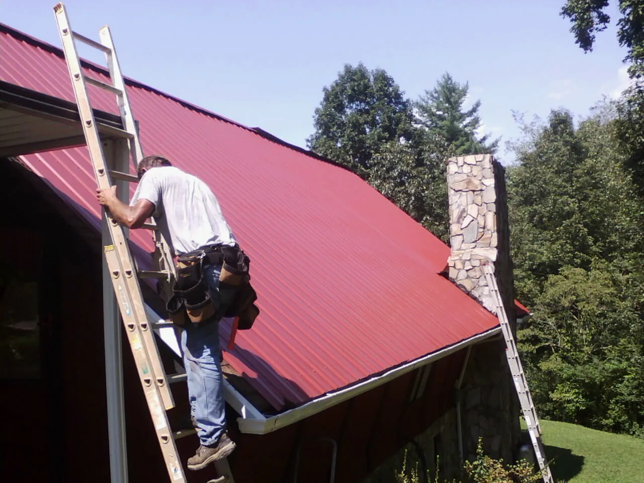 Roofer on a ladder beside a red metal roof and stone chimney — Soffit & Fascia Repair by Roofing Brothers in Pottersdale